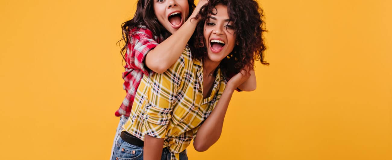 Crazy emotional girls are dabbling and having fun on orange background. Woman laughs and plays hair of her mulatto girlfriend.