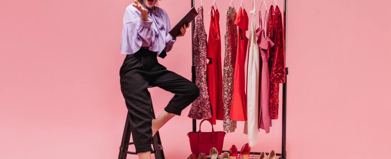 Girl in purple blouse and black pants is sitting on chair, looking into camera and holding folder against background of hangers with bright dresses.