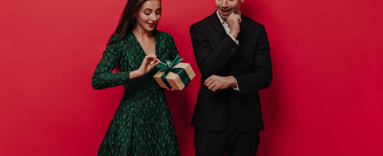handsome-young-man-stylish-black-suit-waiting-his-girlfriend-green-dress-unpack-gift-box-fascinatingly-red-plain-background (1)